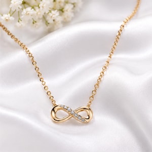 May include: A gold-colored necklace with an infinity symbol pendant. The pendant is adorned with small, clear gemstones. The necklace chain is delicate and the background is a white, silky fabric.
