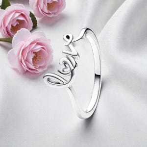 May include: A silver ring with the word "Love" in a cursive font. The ring features a heart-shaped design within the "O" of the word. Pink roses are in the background.