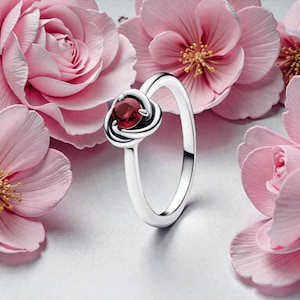 Puede incluir: Un anillo plateado con un diseño de nudo y una gema roja redonda en el centro. El anillo se presenta sobre un fondo de flores rosas, creando una estética romántica y elegante.