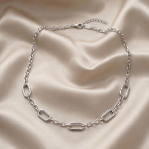May include: A silver-toned chain necklace featuring a series of uniform oval links interspersed with larger, elongated rectangular links. The necklace includes a small circular tag near the lobster claw clasp and an adjustable extender chain.