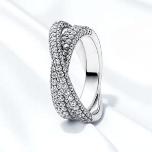 May include: A silver-colored ring with a twisted design. The top portion of the ring is covered in small, sparkling stones, while the band is smooth and polished. The ring is set against a white background.