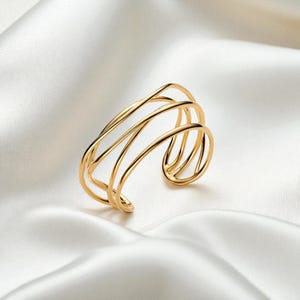 Peut inclure: Un bracelet manchette doré au design moderne et ouvert. Le bracelet présente plusieurs bandes courbes et parallèles qui créent une forme ouverte et fluide. Le bijou est présenté sur une surface blanche semblable à du satin.