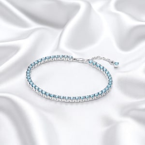 Op de afbeelding: Een zilveren armband met een rij lichtblauwe edelstenen. De armband ligt op een witte, zijdeachtige achtergrond. De sluiting is zichtbaar en de armband is verstelbaar.