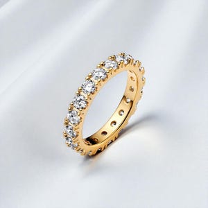 May include: A gold eternity ring featuring a continuous line of clear gemstones. The ring's band has a polished finish, and the gemstones sparkle. The ring is set against a soft, white background.