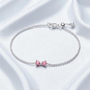 以下が含まれることがあります： 小さなピンクのリボンチャームが付いた繊細なシルバーブレスレット。ブレスレットは細いチェーンのデザインと安全な留め金が付いています。リボンチャームはピンクの宝石で作られています。ブレスレットは白くシルキーな背景に表示されています。