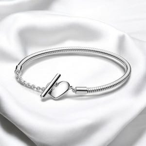 May include: A silver bracelet with a snake chain design. The bracelet features a heart-shaped clasp and a T-bar closure. The jewelry is displayed on a white, silky surface.