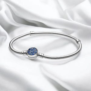 以下が含まれることがあります： 小さな青い宝石がセットされた丸いチャームが付いたシルバーブレスレット。 ブレスレットはスネークチェーンのデザインで、白くシルキーな背景に表示されています。 クラスプは青い石が付いた丸いチャームです。