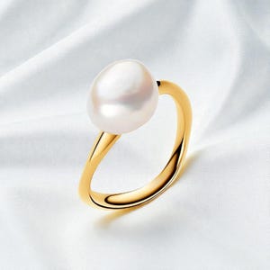 Op de afbeelding: Een gouden ring met een grote, glanzende witte parel. De ring heeft een eenvoudig, elegant ontwerp met een gladde, gepolijste band. De ovaalvormige parel zit prominent op de ring en creëert een klassieke en verfijnde uitstraling.