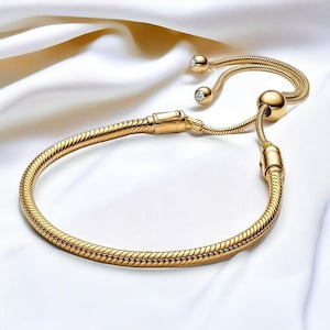 Könnte beinhalten: Ein goldfarbenes Armband mit Schlangenketten-Design. Das Armband verfügt über einen Schiebeverschluss und zwei kleine, runde Anhänger mit klaren Edelsteinen. Das verstellbare Design umfasst eine große, runde Perle und zwei kleinere Perlen.