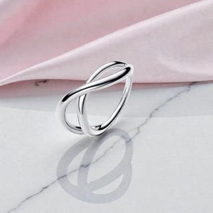 Pode incluir: Um anel de prata polida com um design de símbolo de infinito. O anel apresenta duas bandas entrelaçadas que se cruzam, criando um loop contínuo. O anel está sobre um tecido rosa suave e uma superfície de mármore.