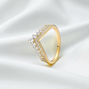 Può includere: Un anello dorato con un design a forma di V. L'anello è adornato da numerose piccole gemme trasparenti sulla parte superiore e sui lati, creando un effetto scintillante. L'anello è posto su uno sfondo bianco.