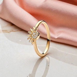 May include: A gold-colored ring featuring a sunburst design. The sun's center is a cluster of small, clear gemstones, with pointed rays extending outward. The ring band is smooth and polished, reflecting light.