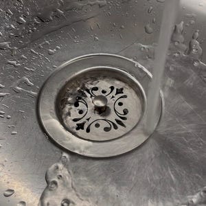 以下が含まれることがあります： 黒いフィリグリー模様が施された円形の排水口を持つステンレス製のキッチンシンク。蛇口からシンクに水が流れ込み、排水口の周りの金属表面に波紋と水滴が広がっています。