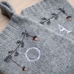 Puede incluir: Dos calcetines navideños de punto gris con detalles bordados. Cada calcetín presenta un lazo para colgar, letras bordadas, ramas de pino y bellotas. Las letras "A" y "O" son visibles en los calcetines.