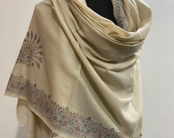 May include: A cream-colored shawl with intricate floral and paisley patterns along the borders and at one corner. The shawl is draped over a black mannequin, showcasing its soft texture and design. The detailed embroidery includes shades of red, green, and brown.