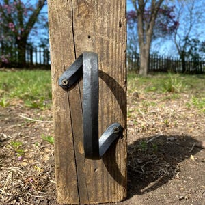 May include: A black metal door handle is attached to a weathered wooden board. The handle is shaped like a curved 'J' and has a rustic, handmade look.