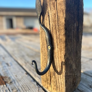 Puede incluir: Un gancho de metal negro con una parte superior en forma de hoja está montado en un bloque de madera desgastado. El gancho está unido al bloque con un pequeño tornillo.