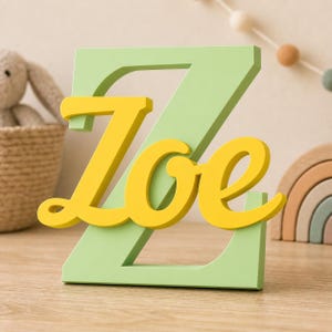 May include: A light green capital letter Z with the name Zoe written in a yellow cursive script font layered over the front. The letter Z is a solid block shape, while the yellow name Zoe features a smooth, flowing, connected typeface.