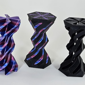 May include: Three 3D printed hourglass-shaped objects with a starburst design on the top. The object on the left is a vibrant purple and blue color, the middle object is black with purple and blue accents, and the object on the right is black.