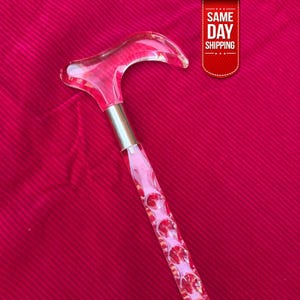 Puede incluir: Un bastón de acrílico rosa transparente con un mango curvo y un collar de metal plateado. El eje presenta una serie de gemas rosas facetadas incrustadas. El bastón está sobre un fondo rojo.