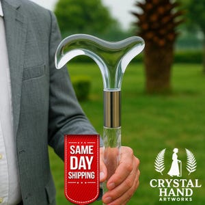 Puede incluir: Un bastón transparente con un mango curvo. El bastón tiene una banda de metal plateado y un eje transparente texturizado. La imagen incluye el texto "Crystal Hand Artworks".