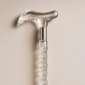 Puede incluir: Un bastón transparente con un mango curvo. El mango y el eje están hechos de material transparente, con un patrón texturizado en el eje. Una banda de metal plateado conecta el mango al eje.