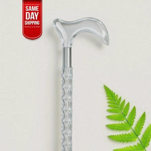 Puede incluir: Un bastón transparente con un mango curvo y un eje texturizado. El bastón tiene una banda de metal plateado que conecta el mango con el eje. Una hoja de helecho verde está en la esquina inferior derecha. Una pancarta roja con el texto "SAME DAY SHIPPING" está en la esquina superior izquierda.