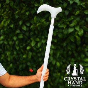 Puede incluir: Un bastón blanco con un mango curvo y un collar de metal plateado. El bastón se sostiene contra un fondo de follaje verde. El logotipo de Crystal Hand Artworks es visible en la esquina inferior derecha.