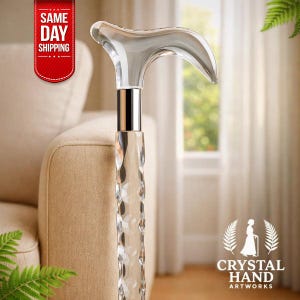 Puede incluir: Bastón de cristal transparente con un mango curvo y una banda de metal plateado. El eje del bastón tiene un diseño texturizado y facetado. El logotipo "Crystal Hand Artworks" es visible.