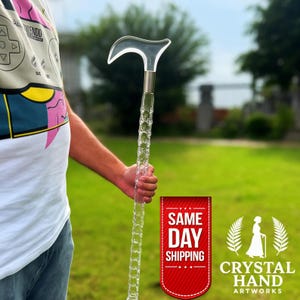 Puede incluir: Un bastón decorativo transparente con un mango curvo y un eje texturizado. El bastón se sostiene frente a un fondo de hierba verde. El bastón tiene un conector de metal plateado. La imagen también incluye una pancarta roja que dice "SAME DAY SHIPPING".