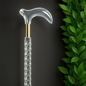 Puede incluir: Un bastón transparente con un mango curvo, un eje texturizado y detalles metálicos dorados. El bastón se encuentra sobre un fondo oscuro con una planta verde y frondosa a la derecha.