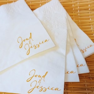 May include: White paper napkins with gold lettering that reads "Joud & Jessica". The napkins are arranged in a fan shape, with the text visible on the top napkin. The background is a woven, light brown surface.