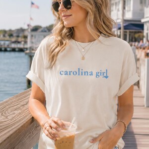 Pu&ograve; includere: Una t-shirt a maniche corte color crema con la scritta "carolina girl" stampata in lettere azzurre sul petto, in cui la lettera "l" &egrave; stilizzata come il contorno dello stato della Carolina del Nord.
