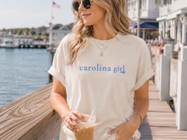 Pu&ograve; includere: Una t-shirt a maniche corte color crema con la scritta "carolina girl" stampata in lettere azzurre sul petto, in cui la lettera "l" &egrave; stilizzata come il contorno dello stato della Carolina del Nord.