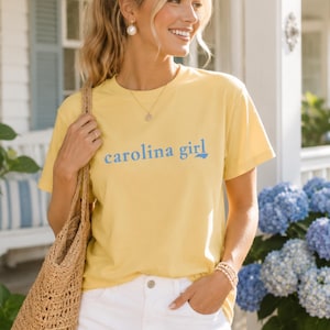 Pu&ograve; includere: Una t-shirt a maniche corte di colore giallo chiaro con la scritta "carolina girl" in lettere blu sul petto. La maglietta presenta un girocollo ed &egrave; abbinata a pantaloni bianchi e a una borsa tote intrecciata color marrone chiaro.
