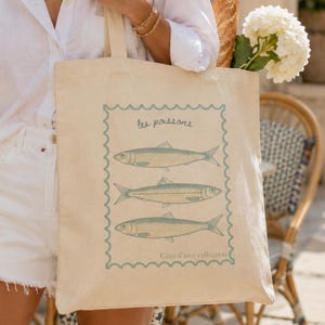 May include: Beige canvas tote bag featuring a blue and white illustration of three fish, with the text "les poissons" and "C&ocirc;te d'azur collection". The bag is held by a person wearing a white shirt and shorts.