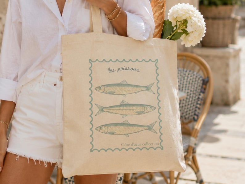 May include: Beige canvas tote bag featuring a blue and white illustration of three fish, with the text "les poissons" and "C&ocirc;te d'azur collection". The bag is held by a person wearing a white shirt and shorts.