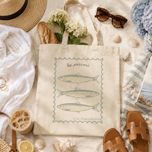 May include: A beige tote bag with a fish illustration, sunglasses, a straw hat, and a book titled "A Summer in the South of France" are arranged on a sandy surface. The bag has the text "les poissons" and "C&ocirc;te d'azur collection".