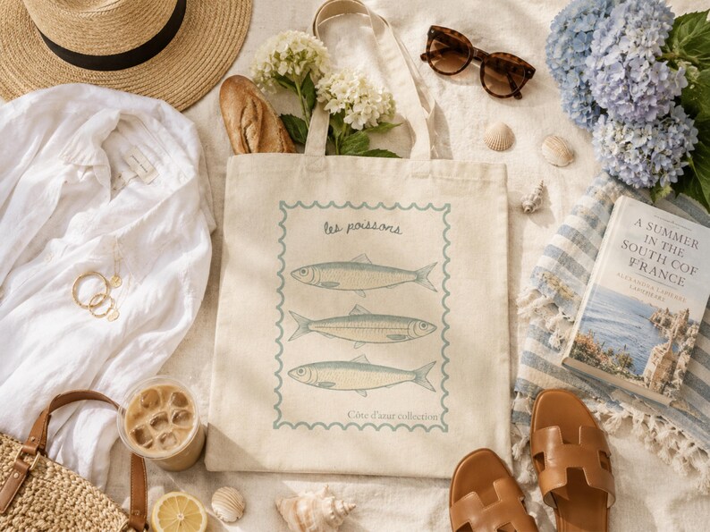 May include: A beige tote bag with a fish illustration, sunglasses, a straw hat, and a book titled "A Summer in the South of France" are arranged on a sandy surface. The bag has the text "les poissons" and "C&ocirc;te d'azur collection".