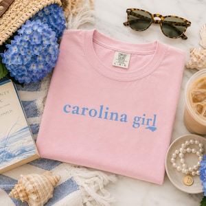 Pu&ograve; includere: Una t-shirt rosa chiaro con la scritta "carolina girl" stampata in blu sul petto. Il capo presenta un girocollo e un'etichetta del marchio Comfort Colors all'interno del colletto. Il tessuto &egrave; in cotone rosa chiaro a tinta unita.
