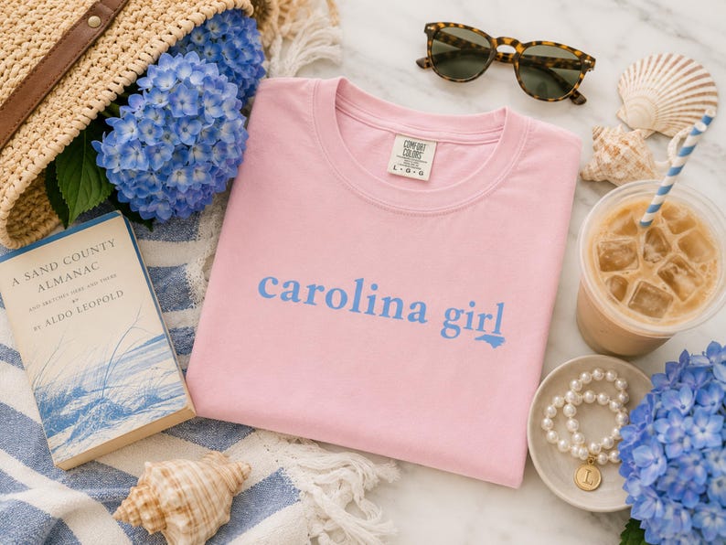 Pu&ograve; includere: Una t-shirt rosa chiaro con la scritta "carolina girl" stampata in blu sul petto. Il capo presenta un girocollo e un'etichetta del marchio Comfort Colors all'interno del colletto. Il tessuto &egrave; in cotone rosa chiaro a tinta unita.
