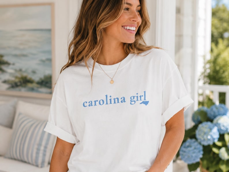 Pu&ograve; includere: Una t-shirt bianca a maniche corte con la scritta "carolina girl" stampata in lettere azzurre sul petto, in cui una piccola sagoma azzurra dello stato della Carolina del Nord sostituisce la lettera "i" nella parola "girl".