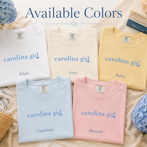 Pu&ograve; includere: Cinque magliette a maniche corte nei colori bianco, avorio, burro, chambray e fiore. Ogni maglietta presenta la scritta 'carolina girl' stampata in blu sul petto. Le magliette sono disposte in gruppo per mostrare le opzioni di colore.
