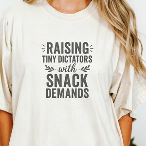 以下が含まれることがあります： 「RAISING TINY DICTATORS with SNACK DEMANDS」のテキストが大胆な濃いグレーのフォントでプリントされた、クリーム色のTシャツ。テキストは中央に配置され、装飾的な要素が施されています。Tシャツは半袖で、ゆったりとしたフィット感です。