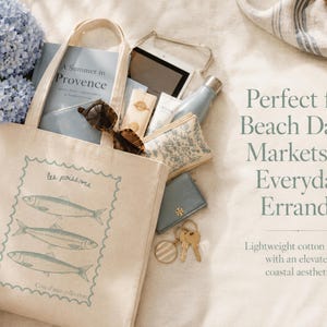 May include: Beige canvas tote bag with a blue fish design, filled with items like sunglasses, a book, and a water bottle. The bag has the text "les poissons" and "A Summer in Provence". Perfect for beach days and errands.