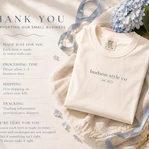 Pu&ograve; includere: Una t-shirt color crema con la scritta 'hudson style co est 2023' stampata in blu scuro sul petto. La maglietta presenta uno scollo rotondo e un'etichetta del marchio Comfort Colors. Il testo a sinistra descrive le politiche aziendali.