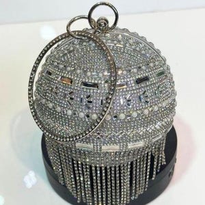 Könnte beinhalten: Eine silberne, perlenbesetzte, kugelförmige Abendtasche mit hängender Fransen. Die Tasche hat einen kreisförmigen Griff und ist mit kleinen, reflektierenden Perlen und Kristallen bedeckt. Die Tasche steht auf einem schwarzen Sockel.