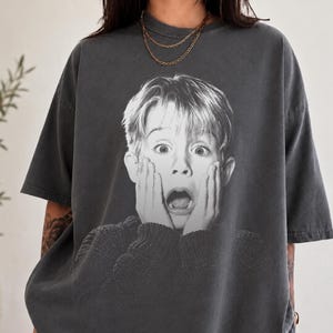 Può includere: Una t-shirt oversize grigio antracite con una grande stampa grafica in bianco e nero di una giovane persona con le mani sulle guance in un'espressione sorpresa, che indossa un maglione lavorato a maglia.