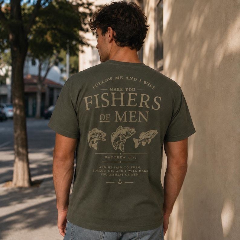 Back view mockup of man wearing olive green Christian fishing t-shirt featuring Fishers of Men Matthew 4:19 Bible verse design with fish illustrations in outdoor urban setting.