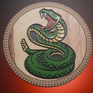 Può includere: Un emblema rotondo in legno raffigurante un serpente verde arrotolato, con la bocca aperta, che mostra una lingua rossa e zanne bianche. Il serpente è posto su uno sfondo marrone chiaro, incorniciato da un bordo decorativo.